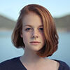 A young woman with red hair standing by a serene body of water.